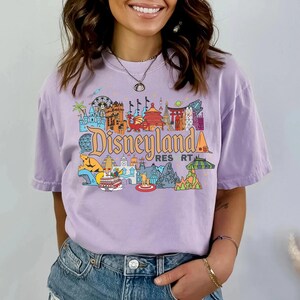 Op de afbeelding: Een lavendelkleurig T-shirt met een kleurrijke Disneyland Resort-afbeelding. Het ontwerp bevat iconische bezienswaardigheden en de tekst Disneyland Resort in een gestileerd lettertype. Het shirt heeft een relaxte pasvorm.