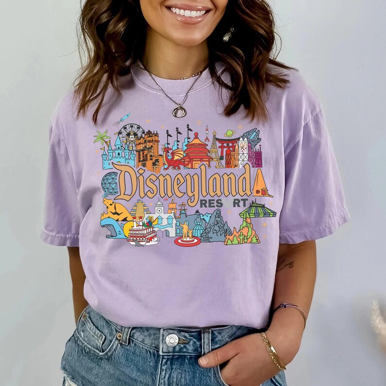 Op de afbeelding: Een lavendelkleurig T-shirt met een kleurrijke Disneyland Resort-afbeelding. Het ontwerp bevat iconische bezienswaardigheden en de tekst Disneyland Resort in een gestileerd lettertype. Het shirt heeft een relaxte pasvorm.