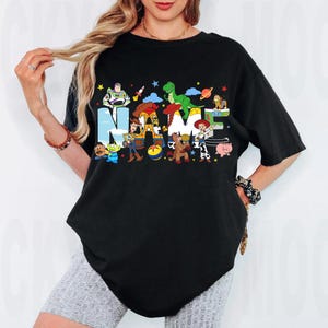 May include: Black t-shirt featuring the word "NAME" in colorful letters with cartoon characters. The design includes characters like Buzz Lightyear, Woody, and Rex. The shirt is a comfortable, oversized fit.