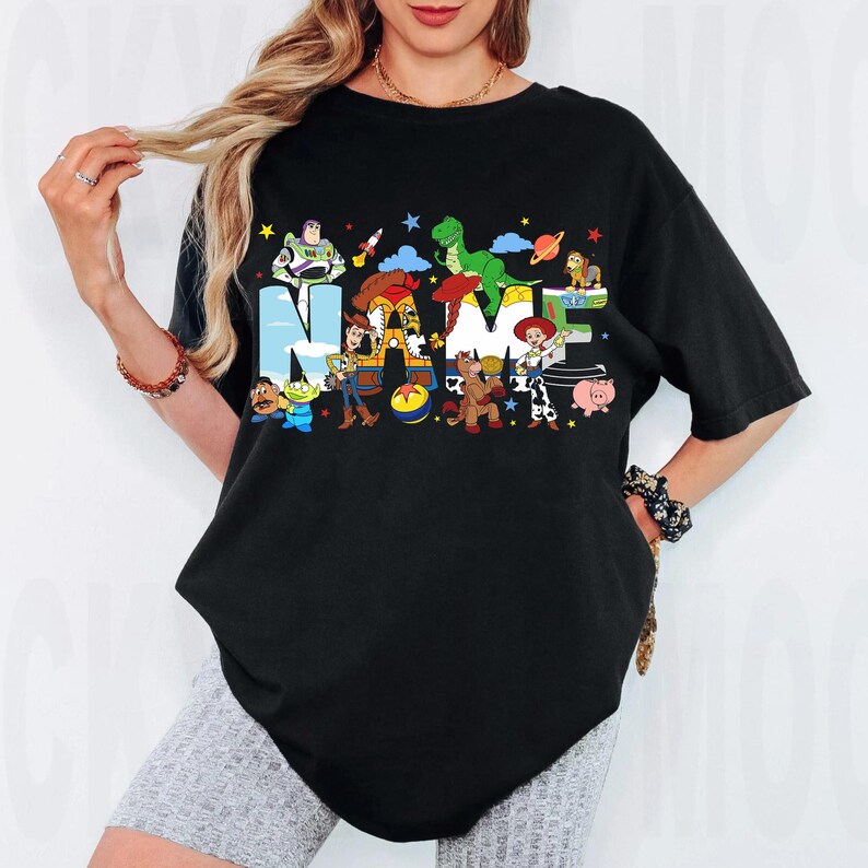 May include: Black t-shirt featuring the word "NAME" in colorful letters with cartoon characters. The design includes characters like Buzz Lightyear, Woody, and Rex. The shirt is a comfortable, oversized fit.