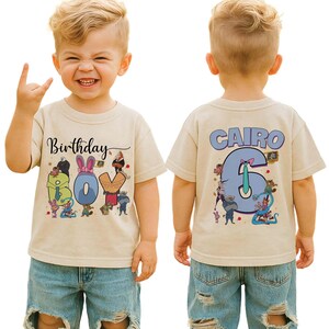 May include: A tan short-sleeved t-shirt with the words "Birthday Boy" and the number "6" printed on it. The front of the shirt has cartoon characters around the words "Boy". The back of the shirt has the name "Cairo" above the number "6".