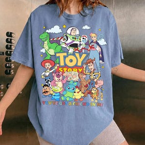 May include: A light blue t-shirt featuring a colorful graphic of characters from the animated film "Toy Story." The design includes the text "TOY STORY" and "YOU'VE GOT A FRIEND IN ME."