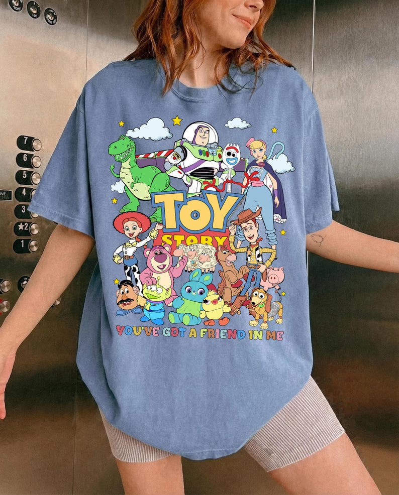 May include: A light blue t-shirt featuring a colorful graphic of characters from the animated film "Toy Story." The design includes the text "TOY STORY" and "YOU'VE GOT A FRIEND IN ME."