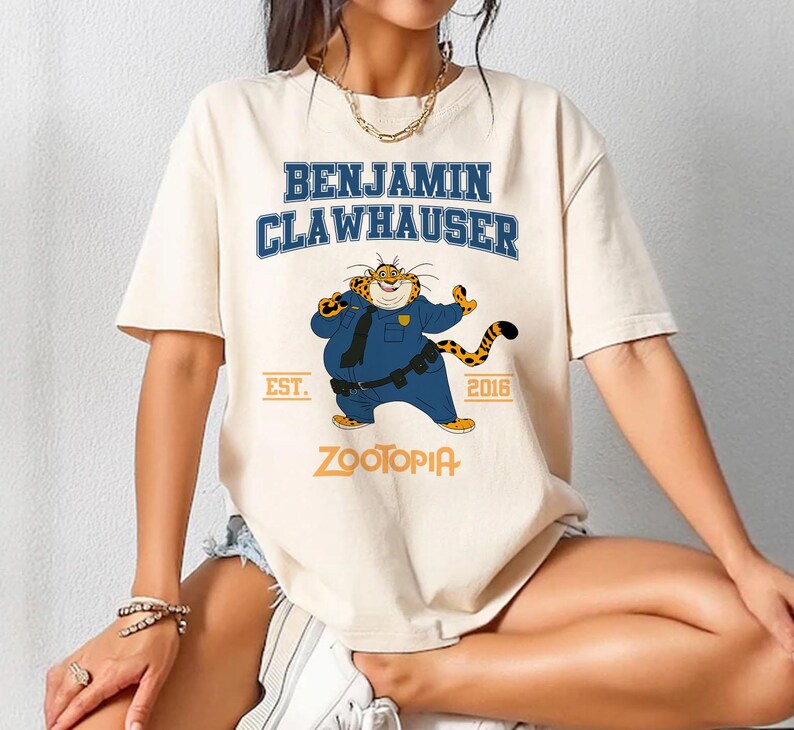 May include: Cream-colored t-shirt featuring the character Benjamin Clawhauser from Zootopia. The design includes the text "BENJAMIN CLAWHAUSER" in blue, the year "2016", and the word "ZOOTOPIA" in orange. The character is depicted in a police uniform.