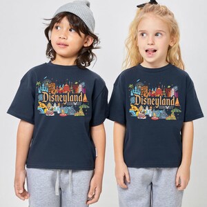 Op de afbeelding: Twee kinderen dragen marineblauwe T-shirts met het Disneyland Resort-logo in kleurrijke graphics. De shirts hebben korte mouwen en een ronde hals. De kinderen dragen grijze joggingbroeken en een grijze beanie.