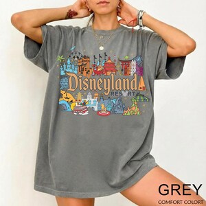 Op de afbeelding: Een grijs t-shirt met een kleurrijke Disneyland Resort-afbeelding. Het ontwerp bevat verschillende parkiconen en het Disneyland-logo. Het shirt heeft korte mouwen en een relaxte pasvorm. Het woord "GREY" staat onderaan gedrukt.