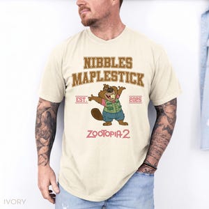 K&ouml;nnte beinhalten: Elfenbeinfarbenes T-Shirt mit einem Cartoon-Biber und dem Text "NIBBLES MAPLESTICK EST. 2025 ZOOTOPIA 2". Der Biber tr&auml;gt eine Weste und Shorts. Das T-Shirt hat kurze &Auml;rmel.