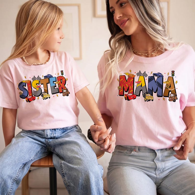 Może przedstawiać: Dwie r&oacute;żowe koszulki z pasującymi wzorami. Jedna koszulka m&oacute;wi "SISTER", a druga "MAMA" w kolorowych, blokowych literach z grafiką samochod&oacute;w wyścigowych. Obie koszulki są połączone z niebieskimi dżinsami.