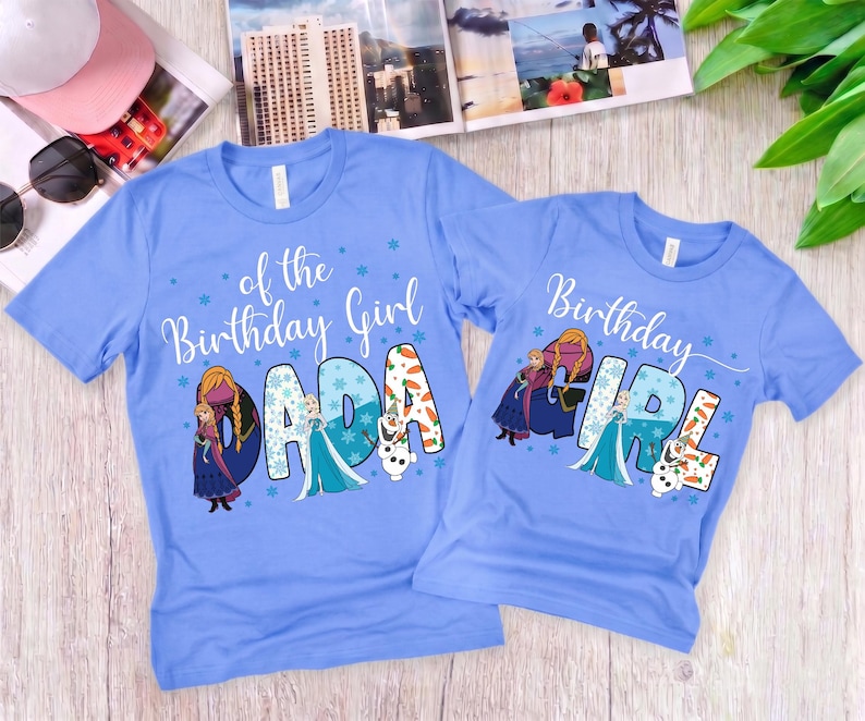 Puede incluir: Dos camisetas azul claro con los personajes de Frozen, Anna, Elsa y Olaf. La camiseta m&aacute;s grande muestra el texto of the Birthday Girl DADA, mientras que la m&aacute;s peque&ntilde;a muestra Birthday GIRL, ambas decoradas con copos y zanahorias.