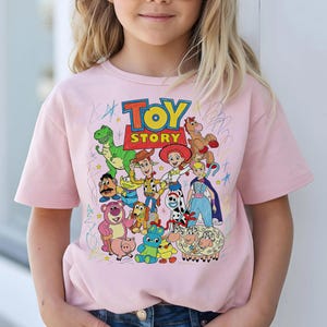 May include: A light pink t-shirt featuring the words "Toy Story" in a colorful graphic. The shirt showcases various cartoon characters from the movie, including Woody, Buzz Lightyear, and others. The design is surrounded by scribbled lines.