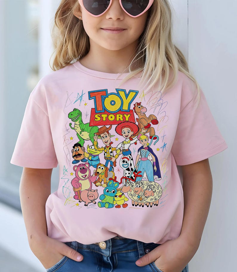 May include: A light pink t-shirt featuring the words "Toy Story" in a colorful graphic. The shirt showcases various cartoon characters from the movie, including Woody, Buzz Lightyear, and others. The design is surrounded by scribbled lines.