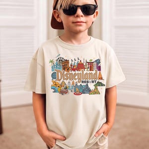 Op de afbeelding: Een beige t-shirt met een kleurrijke afbeelding van Disneyland Resort. De afbeelding bevat verschillende bezienswaardigheden en het Disneyland Resort-logo in een gestileerd lettertype. Het shirt heeft korte mouwen.