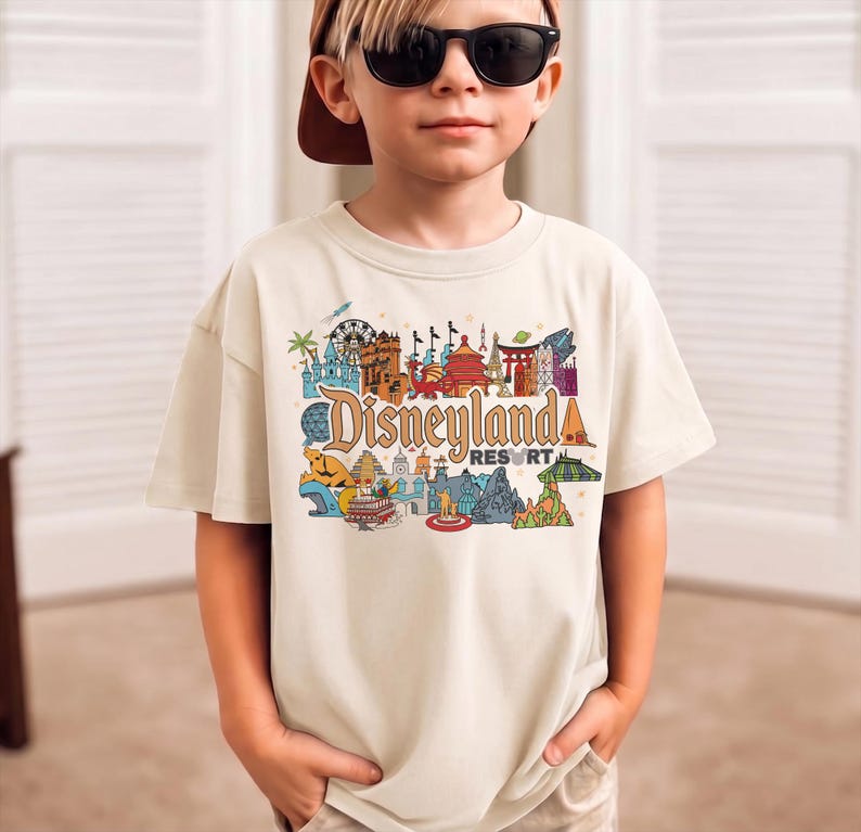 Op de afbeelding: Een beige t-shirt met een kleurrijke afbeelding van Disneyland Resort. De afbeelding bevat verschillende bezienswaardigheden en het Disneyland Resort-logo in een gestileerd lettertype. Het shirt heeft korte mouwen.