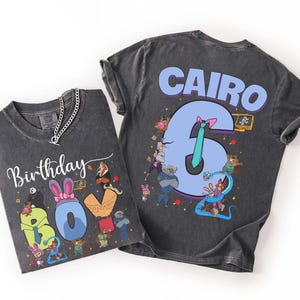 May include: Two dark gray t-shirts with colorful cartoon graphics. One shirt says "Birthday Boy" with a cartoon design. The other shirt says "Cairo 6" with a cartoon design. Both shirts have a chain necklace.