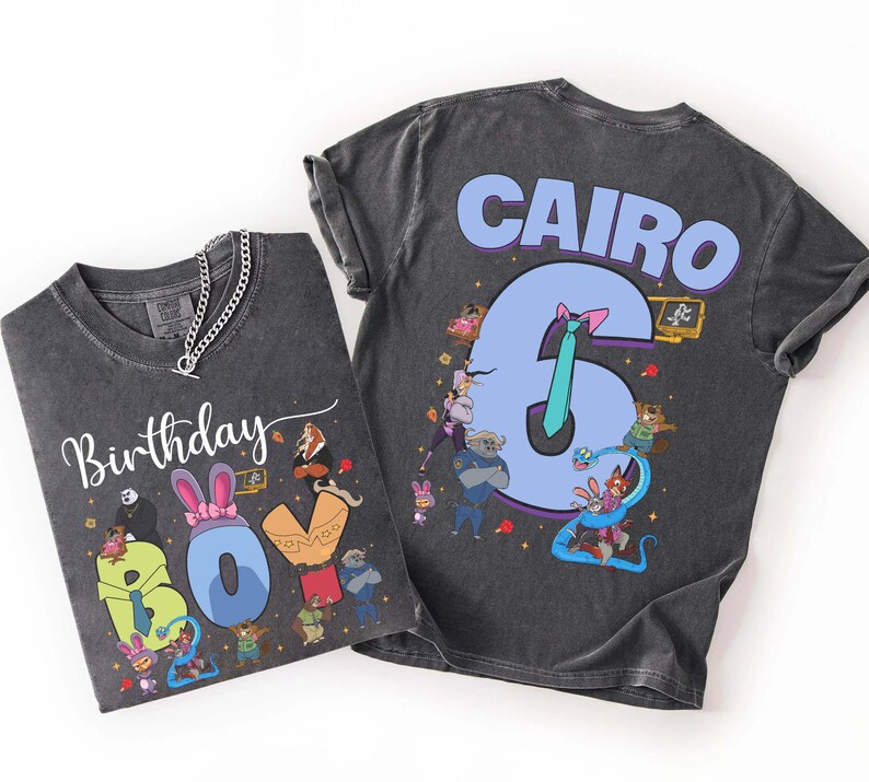 May include: Two dark gray t-shirts with colorful cartoon graphics. One shirt says "Birthday Boy" with a cartoon design. The other shirt says "Cairo 6" with a cartoon design. Both shirts have a chain necklace.