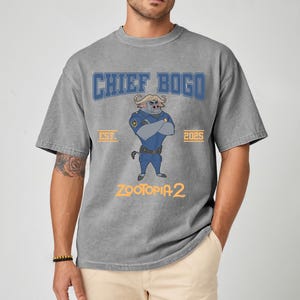 K&ouml;nnte beinhalten: Graues T-Shirt mit Chief Bogo aus Zootopia 2. Das Design zeigt den Schriftzug "CHIEF BOGO" in Blau, die Charakterillustration und den Text "ZOOTOPIA 2" in Gelb. Das Shirt hat auch den Aufdruck "EST. 2025".