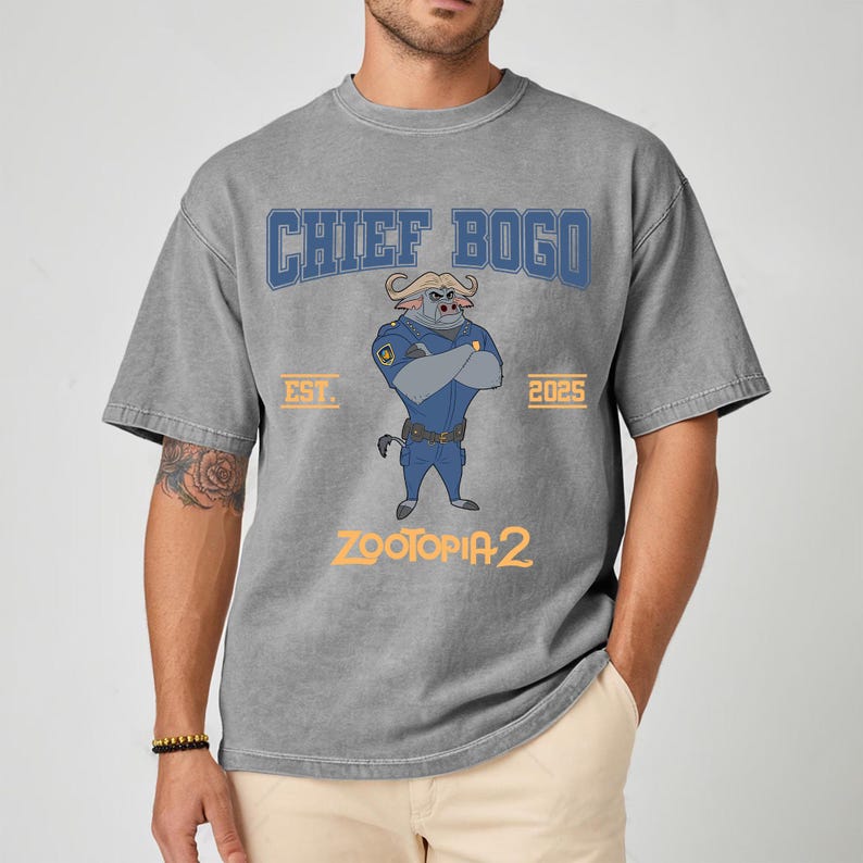 K&ouml;nnte beinhalten: Graues T-Shirt mit Chief Bogo aus Zootopia 2. Das Design zeigt den Schriftzug "CHIEF BOGO" in Blau, die Charakterillustration und den Text "ZOOTOPIA 2" in Gelb. Das Shirt hat auch den Aufdruck "EST. 2025".