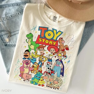 May include: Ivory-colored crew neck t-shirt featuring a colorful graphic of Toy Story characters. The design includes Woody, Buzz Lightyear, and other characters from the animated film. The shirt is paired with blue jeans and a necklace.
