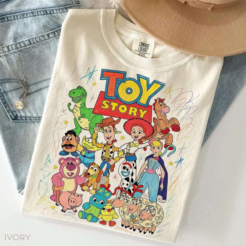 May include: Ivory-colored crew neck t-shirt featuring a colorful graphic of Toy Story characters. The design includes Woody, Buzz Lightyear, and other characters from the animated film. The shirt is paired with blue jeans and a necklace.