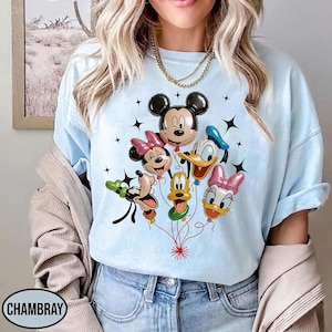 May include: Light blue t-shirt featuring a graphic of Mickey Mouse, Minnie Mouse, Donald Duck, Daisy Duck, Goofy, and Pluto as balloon characters. The balloons are connected by red strings, with black star accents. The shirt has the word "CHAMBRAY" printed on it.