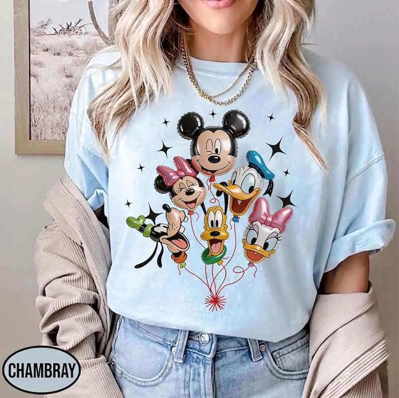 May include: Light blue t-shirt featuring a graphic of Mickey Mouse, Minnie Mouse, Donald Duck, Daisy Duck, Goofy, and Pluto as balloon characters. The balloons are connected by red strings, with black star accents. The shirt has the word "CHAMBRAY" printed on it.