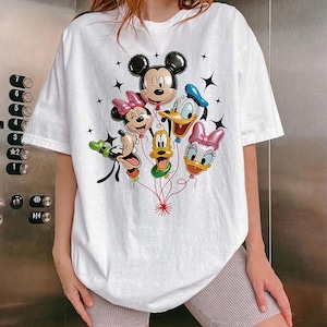 May include: White t-shirt featuring a colorful graphic of Mickey Mouse, Minnie Mouse, Donald Duck, Daisy Duck, Goofy, and Pluto as balloon characters. The design includes black star accents and red string details.