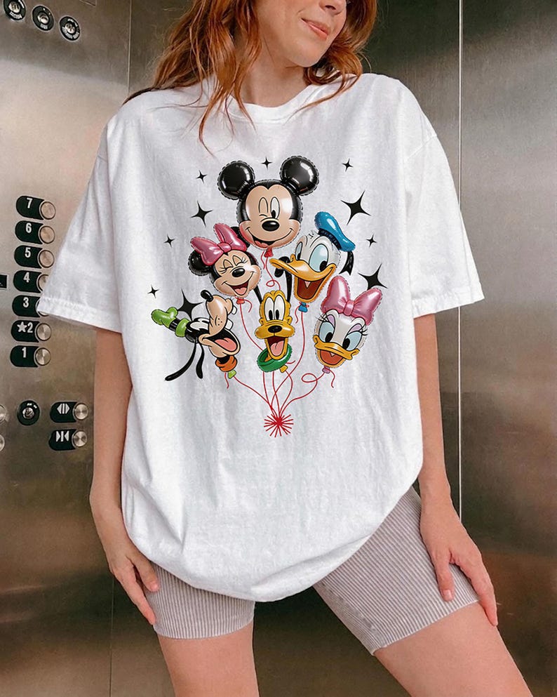 May include: White t-shirt featuring a colorful graphic of Mickey Mouse, Minnie Mouse, Donald Duck, Daisy Duck, Goofy, and Pluto as balloon characters. The design includes black star accents and red string details.