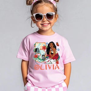 May include: A pink t-shirt featuring a graphic of a cartoon character, flowers, and the number 5. The name "OLIVIA" is printed in orange script. The shirt is worn by a young girl wearing sunglasses and a pink headband.