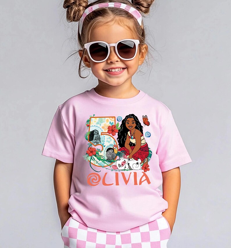 May include: A pink t-shirt featuring a graphic of a cartoon character, flowers, and the number 5. The name "OLIVIA" is printed in orange script. The shirt is worn by a young girl wearing sunglasses and a pink headband.