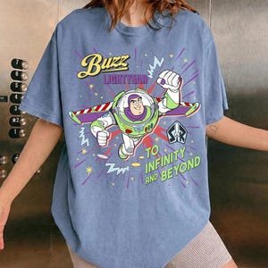 May include: A pale blue t-shirt with a graphic of Buzz Lightyear. The design includes the text "Buzz Lightyear" and "To Infinity and Beyond". The cartoon image shows Buzz in a flying pose.