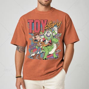 May include: Rust-colored t-shirt featuring a graphic of Toy Story characters Woody, Buzz Lightyear, and Rex. The text "TOY STORY" and "SINCE 1995" are also displayed. The shirt has a vintage, distressed look.