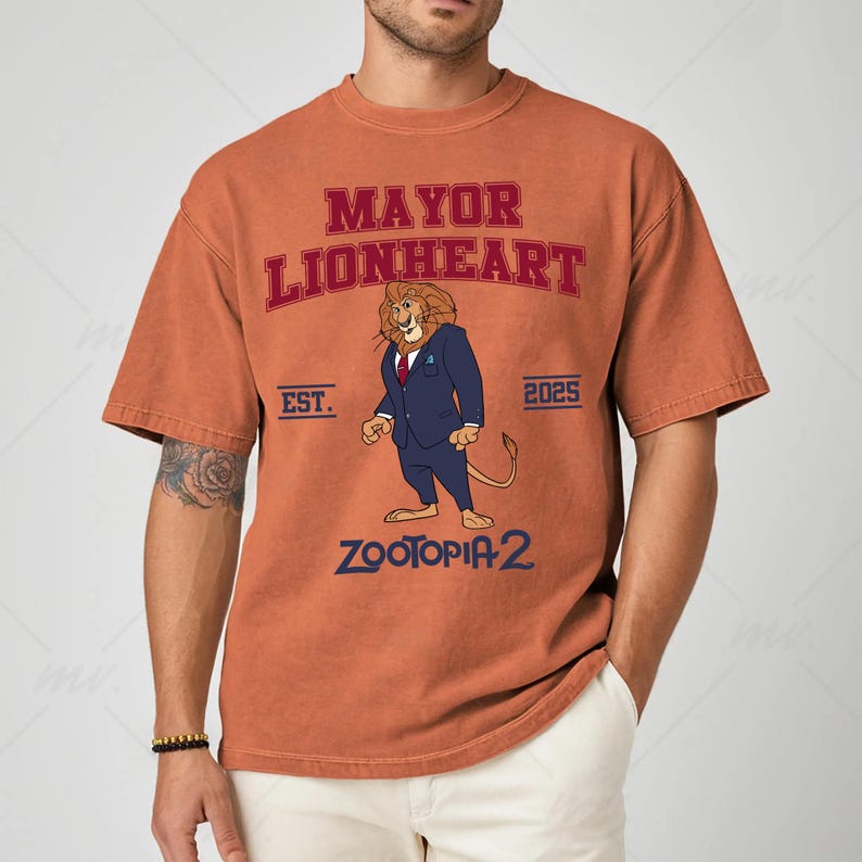 K&ouml;nnte beinhalten: Orangebraunes T-Shirt mit einem Cartoon-L&ouml;wen im Anzug, dem Text "MAYOR LIONHEART" dar&uuml;ber und "ZOOTOPIA 2" darunter. Das Shirt enth&auml;lt auch "EST. 2025".