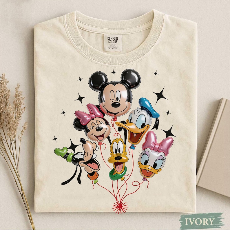 May include: Ivory-colored t-shirt featuring a graphic of Mickey Mouse, Minnie Mouse, Donald Duck, Daisy Duck, and Goofy as balloon characters. The balloons are connected by red strings, with black star accents. The shirt has a crew neck and short sleeves.