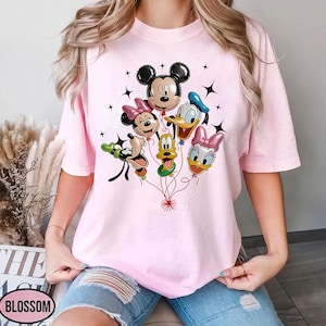 May include: A light pink t-shirt featuring a graphic of Mickey Mouse, Minnie Mouse, Donald Duck, Daisy Duck, and Goofy as balloon characters. The balloons are connected by red strings, with black star accents. The word "BLOSSOM" is visible.
