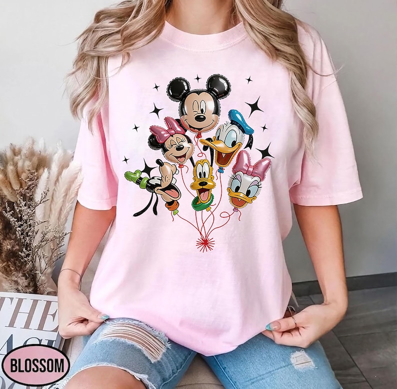 May include: A light pink t-shirt featuring a graphic of Mickey Mouse, Minnie Mouse, Donald Duck, Daisy Duck, and Goofy as balloon characters. The balloons are connected by red strings, with black star accents. The word "BLOSSOM" is visible.