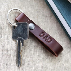 Könnte beinhalten: Ein brauner Leder-Schlüsselanhänger mit dem geprägten Wort "DAD" und einem silbernen Schlüsselring. Ein silberner Schlüssel ist befestigt. Der Schlüsselanhänger liegt auf einer beigen Oberfläche neben einem Stapel blauer Bücher.