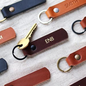 May include: An assortment of personalized leather keychains in various colors, including brown, black, and tan. Some keychains have names or initials embossed in gold lettering. A gold key is attached to one keychain.