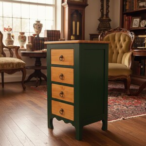 May include: A dark green wooden cabinet with four drawers. The drawer fronts are light wood, and each has a dark metal pull. The top of the cabinet is also light wood. The cabinet is in a room with a window and antique furniture.
