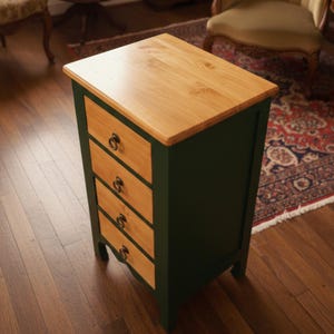 May include: A small, dark green wooden cabinet with four drawers and a light wood top. The drawers have brass ring pulls. The cabinet is a classic piece of furniture, suitable for a bedroom or living room.
