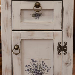 May include: A distressed white cabinet with a drawer and a door. The drawer and door feature floral decals and brass ring pulls. The cabinet has a rustic, vintage aesthetic, with visible wood grain and distressed paint.