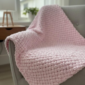 May include: A soft, pink woven blanket draped over a gray armchair. The blanket has a textured, basket-weave pattern. The chair is in front of a window with a lamp and side table visible in the background.