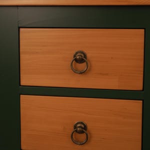 May include: Close-up of a wooden cabinet with two drawers. The drawers are light brown with a visible wood grain. Each drawer has a circular, antique-style metal handle. The cabinet frame is dark green.