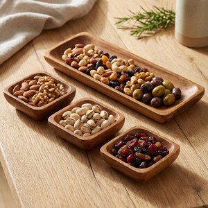 Può includere: Un vassoio di legno e quattro piccole ciotole di legno piene di snack. Il vassoio rettangolare contiene un mix di noci, olive e frutta secca. Le ciotole quadrate contengono mandorle, noci, pistacchi e mirtilli rossi secchi. Gli oggetti in legno sono su una superficie di legno.