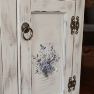 May include: A distressed white cabinet door with a floral design in shades of purple and green. The door features a round, antique brass handle and hinges. The cabinet has a rustic, farmhouse style.