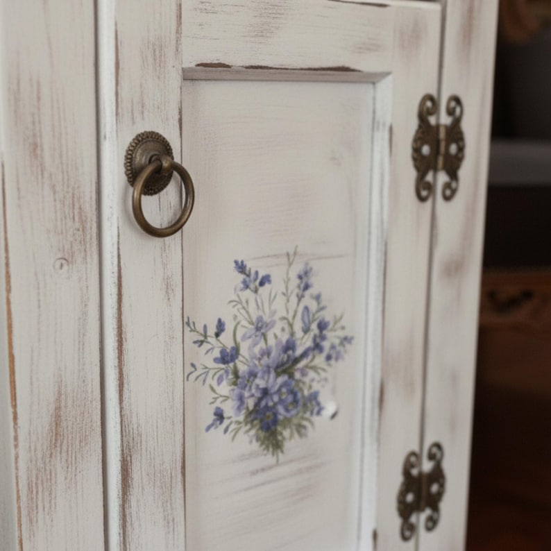 May include: A distressed white cabinet door with a floral design in shades of purple and green. The door features a round, antique brass handle and hinges. The cabinet has a rustic, farmhouse style.