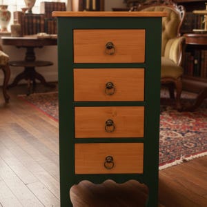 May include: A dark green wooden cabinet with four drawers. The drawer fronts are light brown with brass ring pulls. The top of the cabinet is light brown. The cabinet has a decorative base and is in a room with antique furniture.