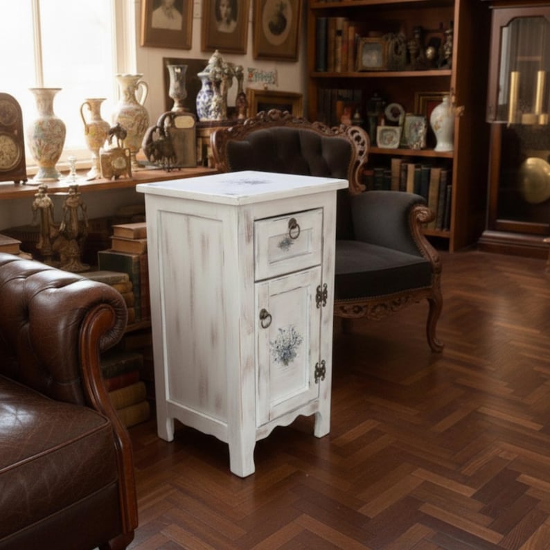 May include: A distressed white wooden bedside cabinet with a drawer and a door. The cabinet features dark hardware and a floral design on the door. The cabinet is in a room with antique furniture and decor.