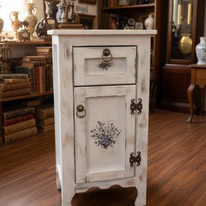 May include: A distressed white wooden nightstand with a drawer and cabinet door. The drawer and door feature floral designs and antique brass hardware. The nightstand has a rustic, vintage aesthetic, perfect for a bedroom or living room.