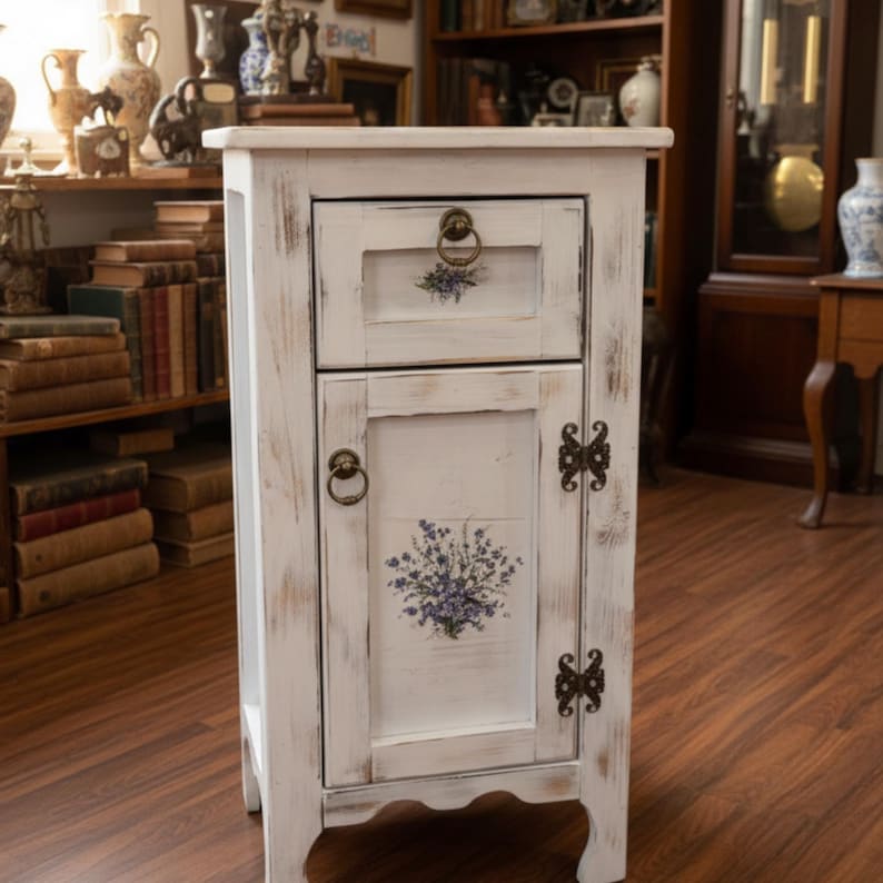 May include: A distressed white wooden nightstand with a drawer and cabinet door. The drawer and door feature floral designs and antique brass hardware. The nightstand has a rustic, vintage aesthetic, perfect for a bedroom or living room.