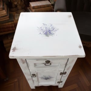 May include: White painted bedside table with a distressed finish. The table features a drawer and a cabinet door, both adorned with a lavender floral design. The top of the table also has a lavender floral design. The hardware is antique brass.